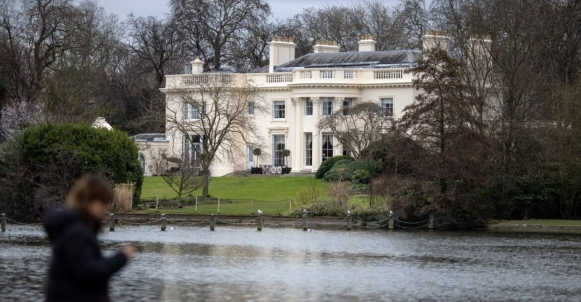 White neoclassical house sits beside a lake with a green lawn and bare trees framing the scene; a blurred person in a dark jacket is in the foreground with a device in hand.