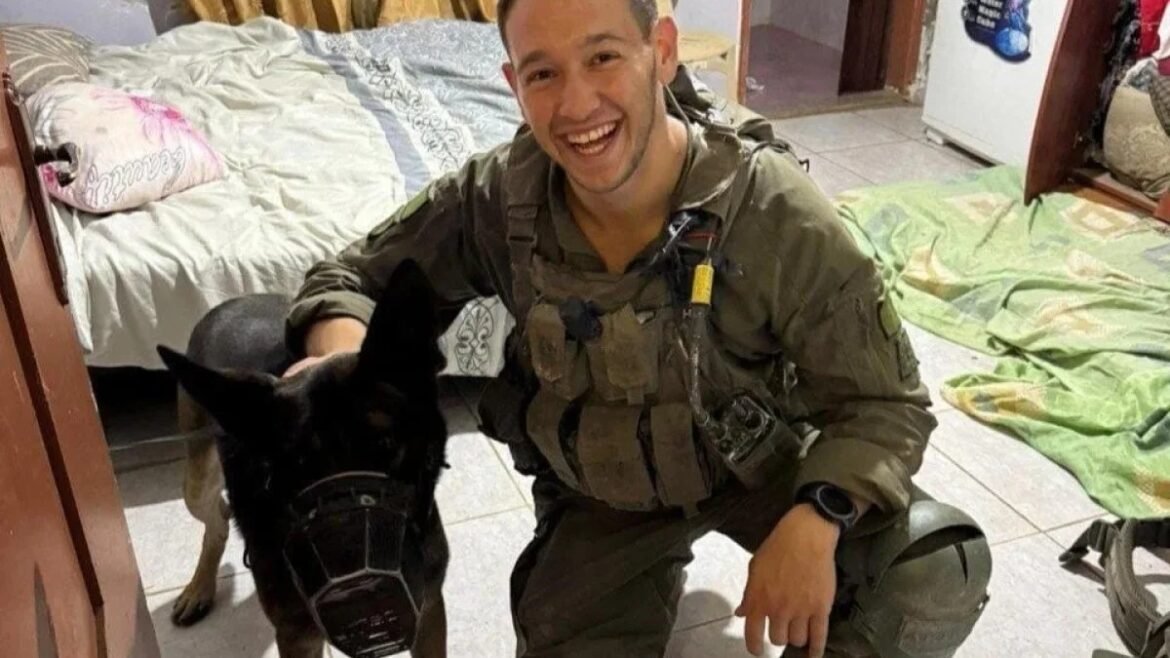 Soldier in tactical gear kneels beside a black dog wearing a muzzle indoors in a bedroom.