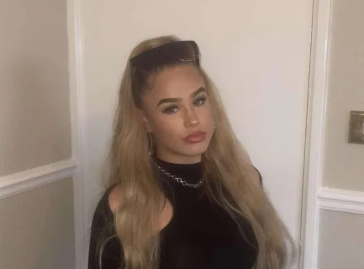 Young woman with long blonde hair, black top, sunglasses on her head, standing in a hallway by a door, gazing slightly to the side.