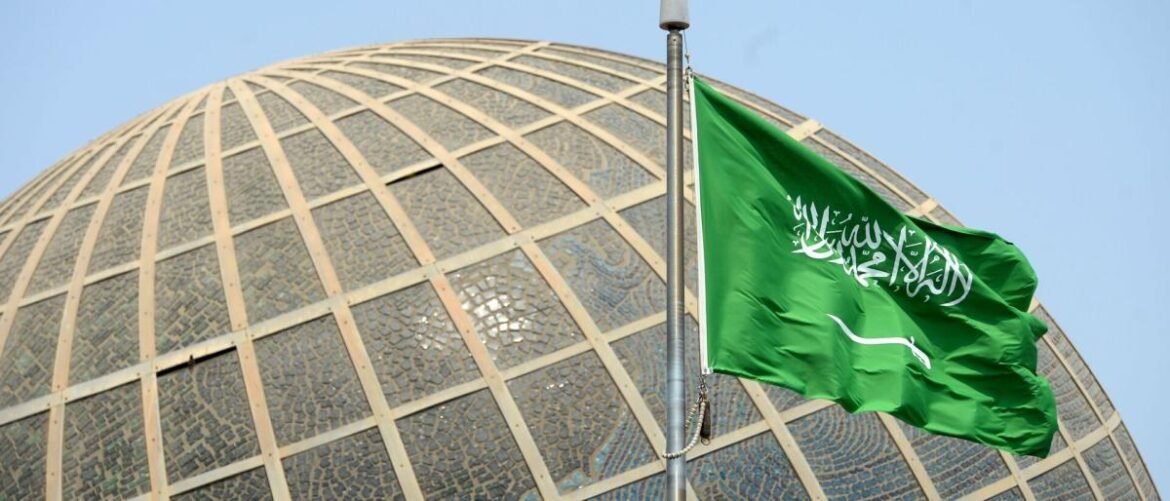Green Saudi Arabia flag waving in front of a glass mosaic dome on a clear day