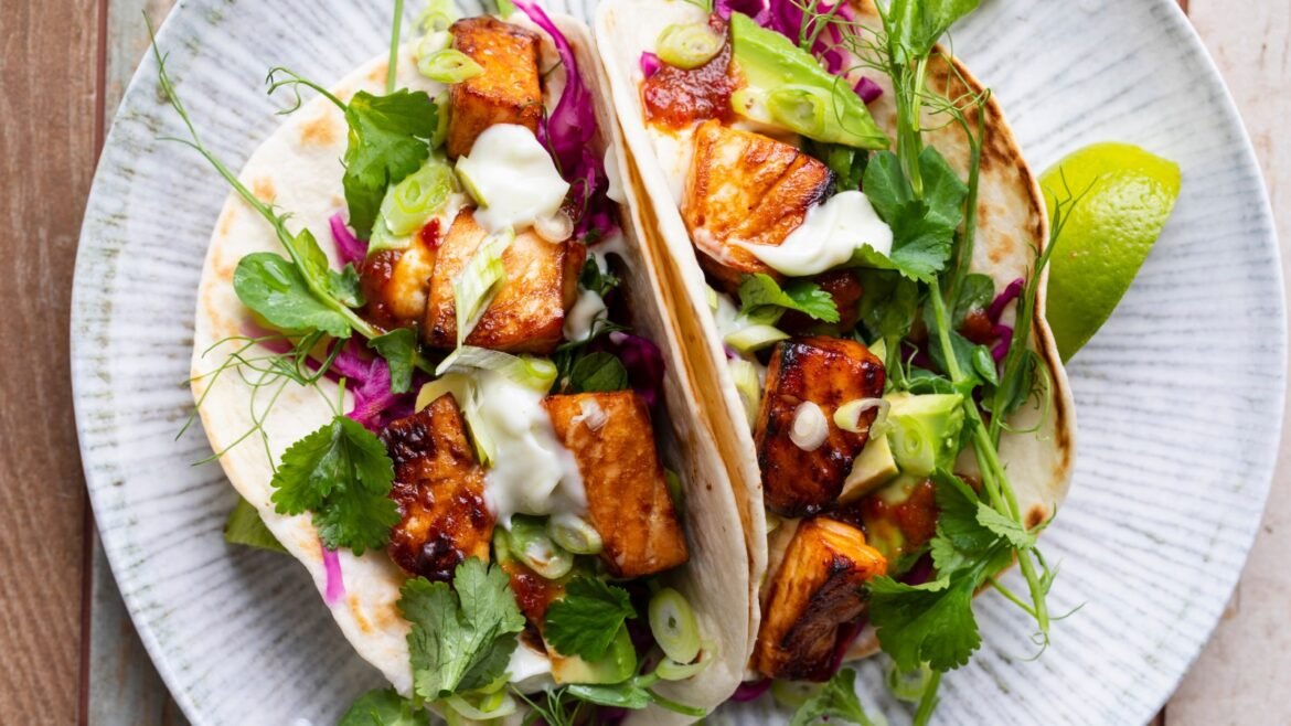 Two soft tacos filled with grilled tofu, fresh greens, cilantro, and a lime wedge on a light plate.