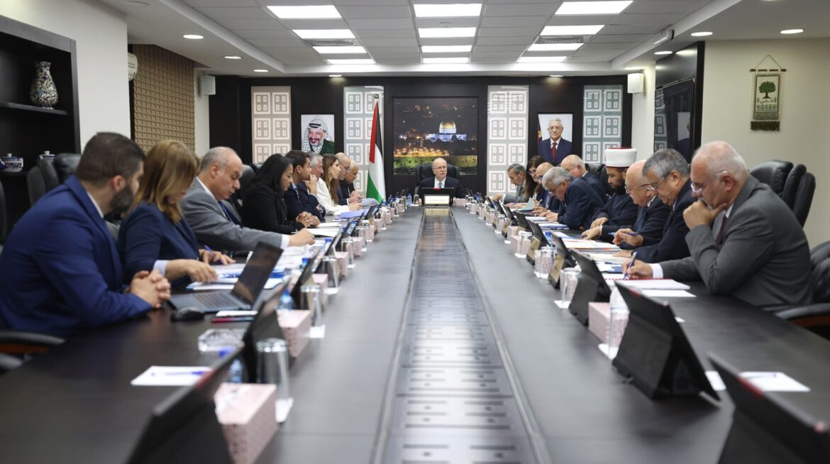 Wide view of a formal boardroom with a long central table, officials in suits seated on both sides, Palestinian flag at the far end, and framed portraits on the back wall behind the speaker.