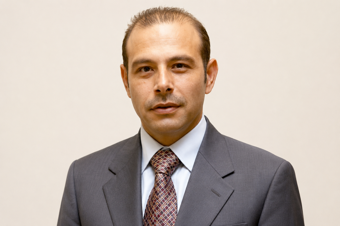 Professional headshot of a man in a dark gray suit and patterned tie facing the camera in a neutral setting.