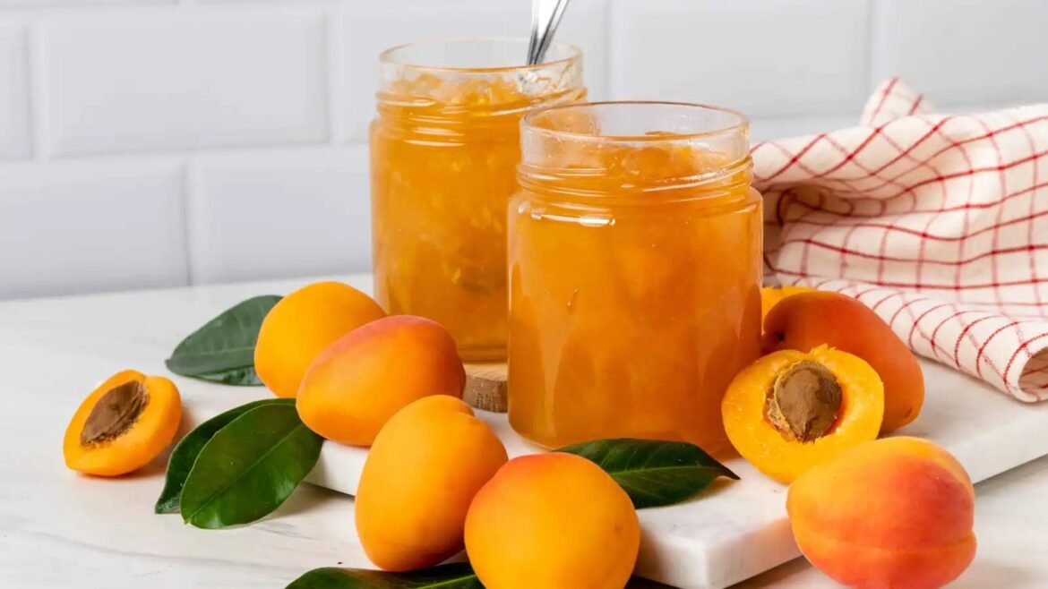 Two glass jars of apricot jam on a white marble surface with fresh apricots and green leaves nearby.