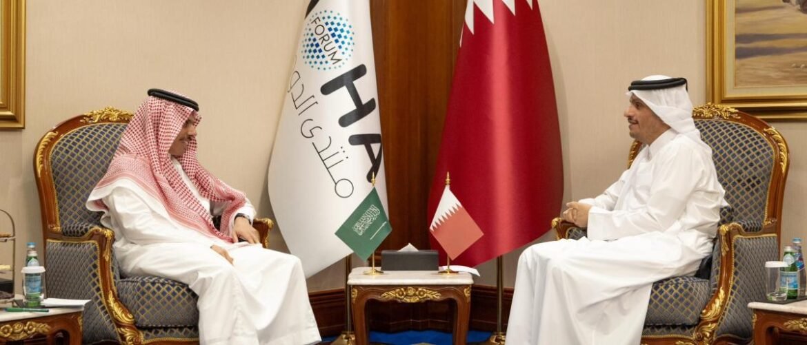 Two men in traditional Middle Eastern attire sit facing each other in ornate chairs during a formal meeting, with flags between them.