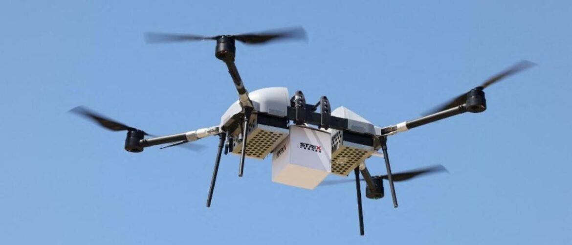 Quadcopter drone in flight carrying a white delivery box beneath it against a clear blue sky (delivery drone).