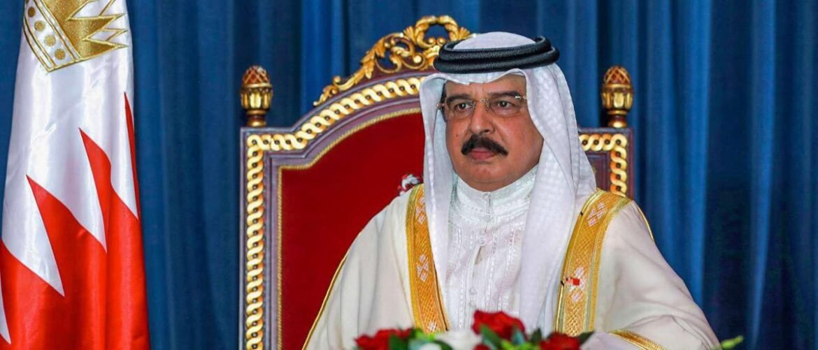 Man in white kandura and head covering sits on an ornate red throne, with a Bahraini flag beside him, during a formal ceremony.