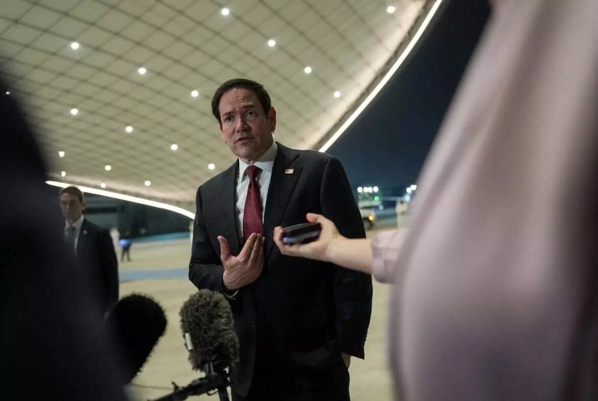 Man in a dark suit being interviewed by reporters at an airport at night, microphones in front of him.