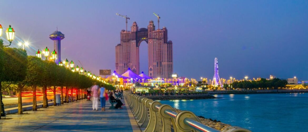 Evening waterfront promenade with illuminated lamps, pedestrians, and a tall arch-shaped building under construction across the water.