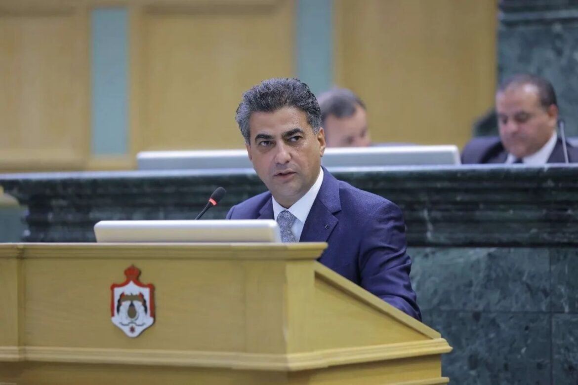 A man in a dark suit speaks at a wooden podium with a crest/emblem on the front, in a formal chamber with microphones nearby.