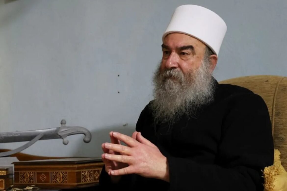 Bearded man in a white clerical hat and black robe sits with hands clasped.