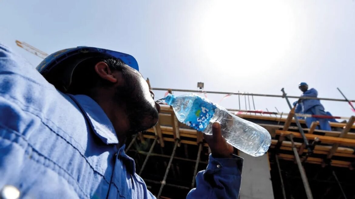 Construction worker in a blue shirt and hardhat drinks water from a plastic bottle at a building site with scaffolding in the background.