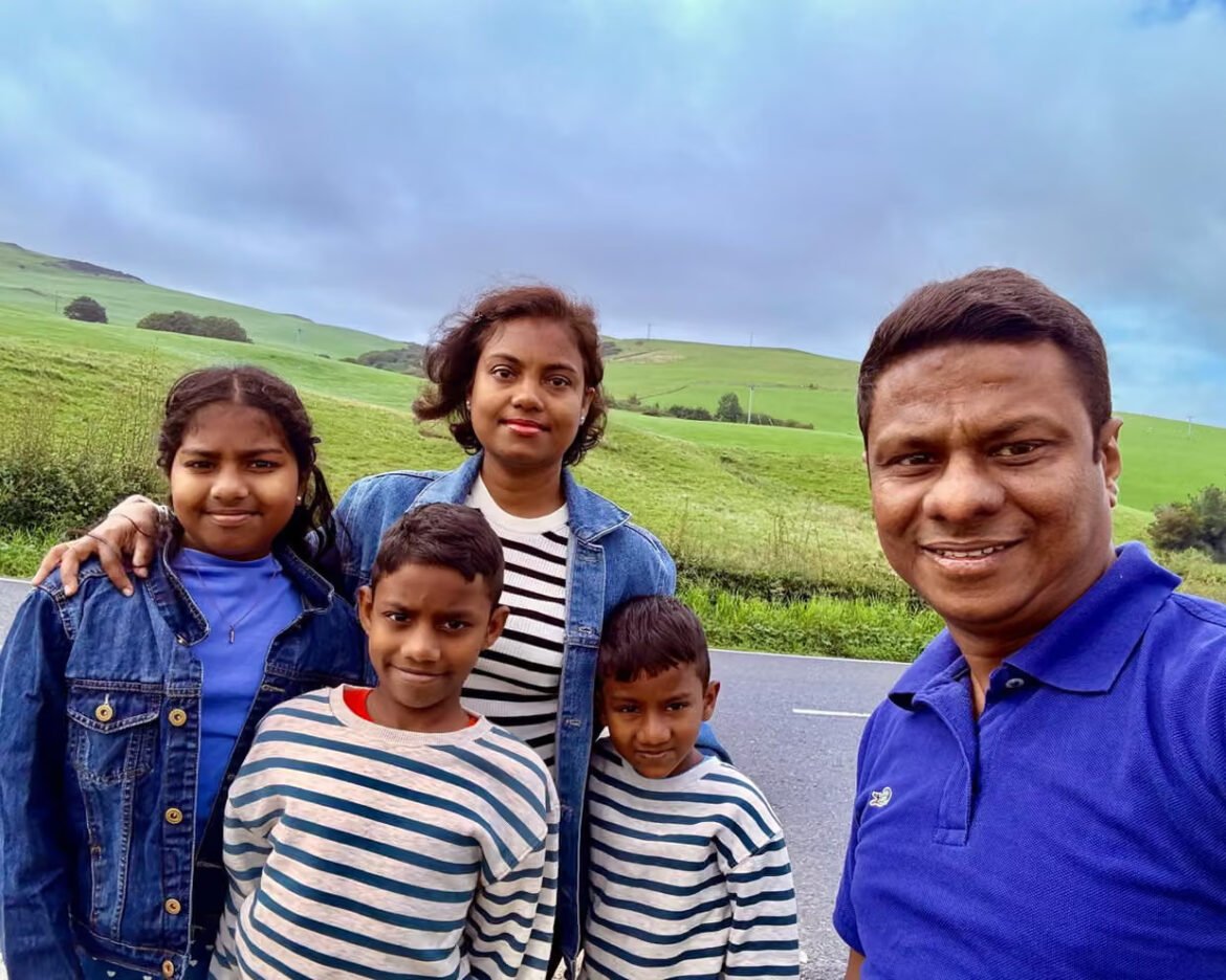 Family of five posing for a selfie on a road beside green rolling fields under a cloudy sky, with two girls in denim jackets and two boys in striped shirts beside a man in a blue polo take the photo.