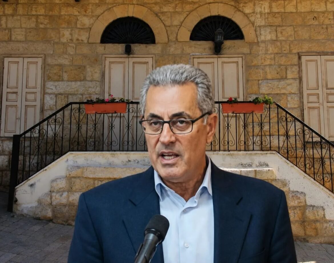 Man in a suit speaks into a microphone outdoors, with a stone building and railing behind him.