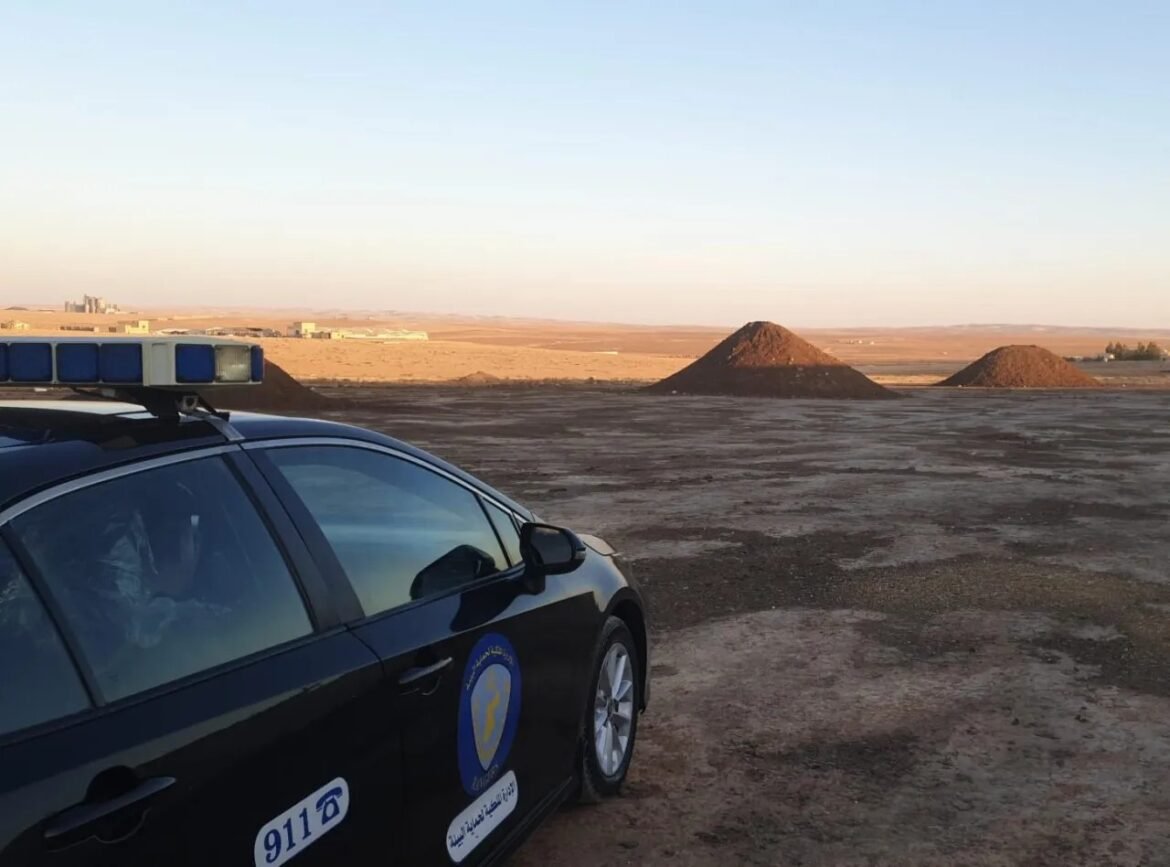 Black police car with a 911 sticker and blue emergency lights on a dusty desert lot, with dirt piles and a distant horizon in view.