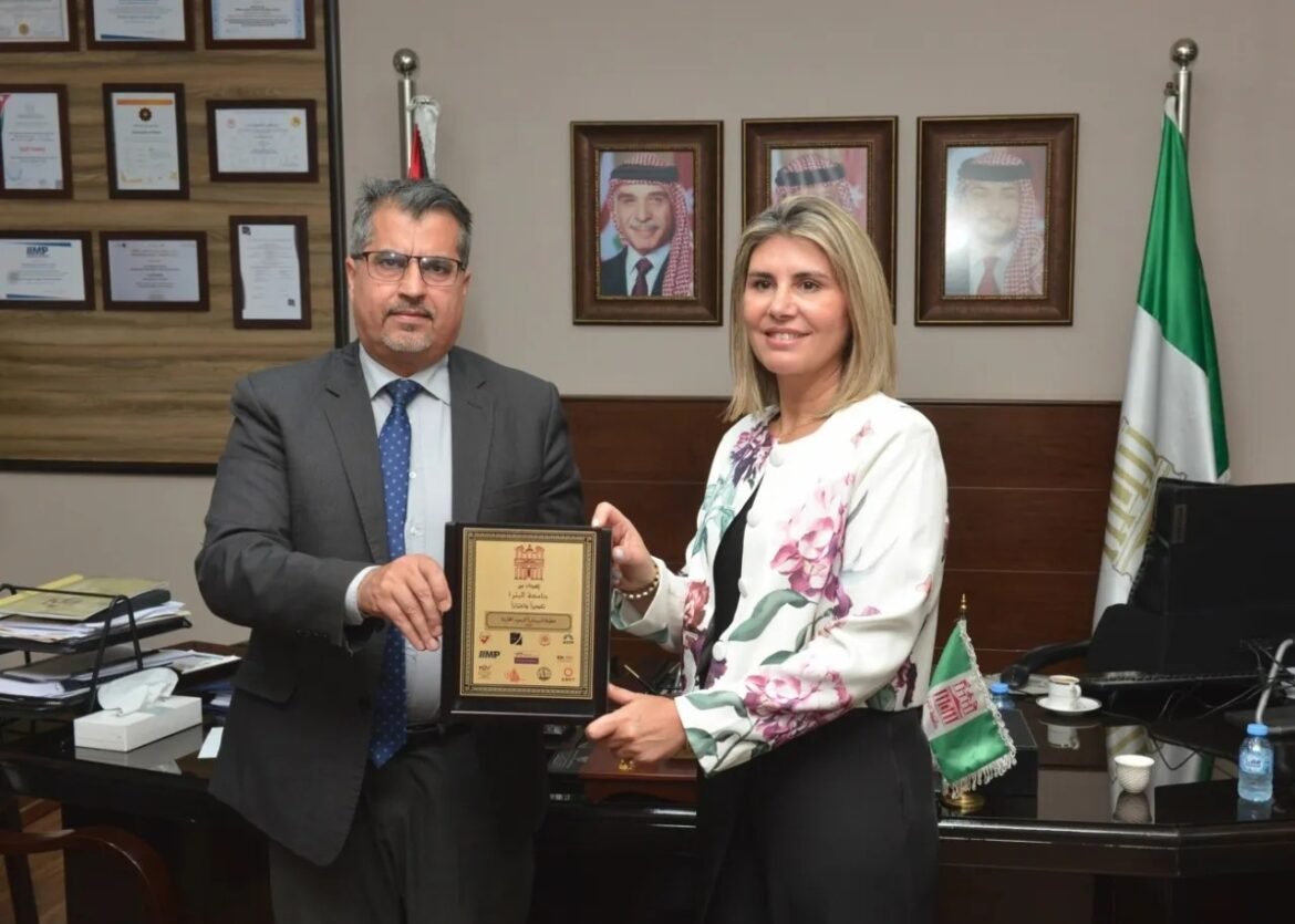 Man in a gray suit and woman in a floral blazer exchange a framed certificate in an office with portraits on the wall and flags in the background.