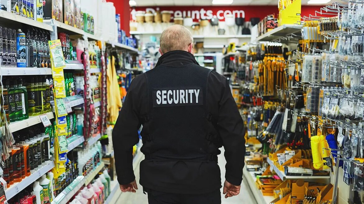 Security guard wearing a black vest with 'SECURITY' on the back, standing in a store aisle facing away from the camera.