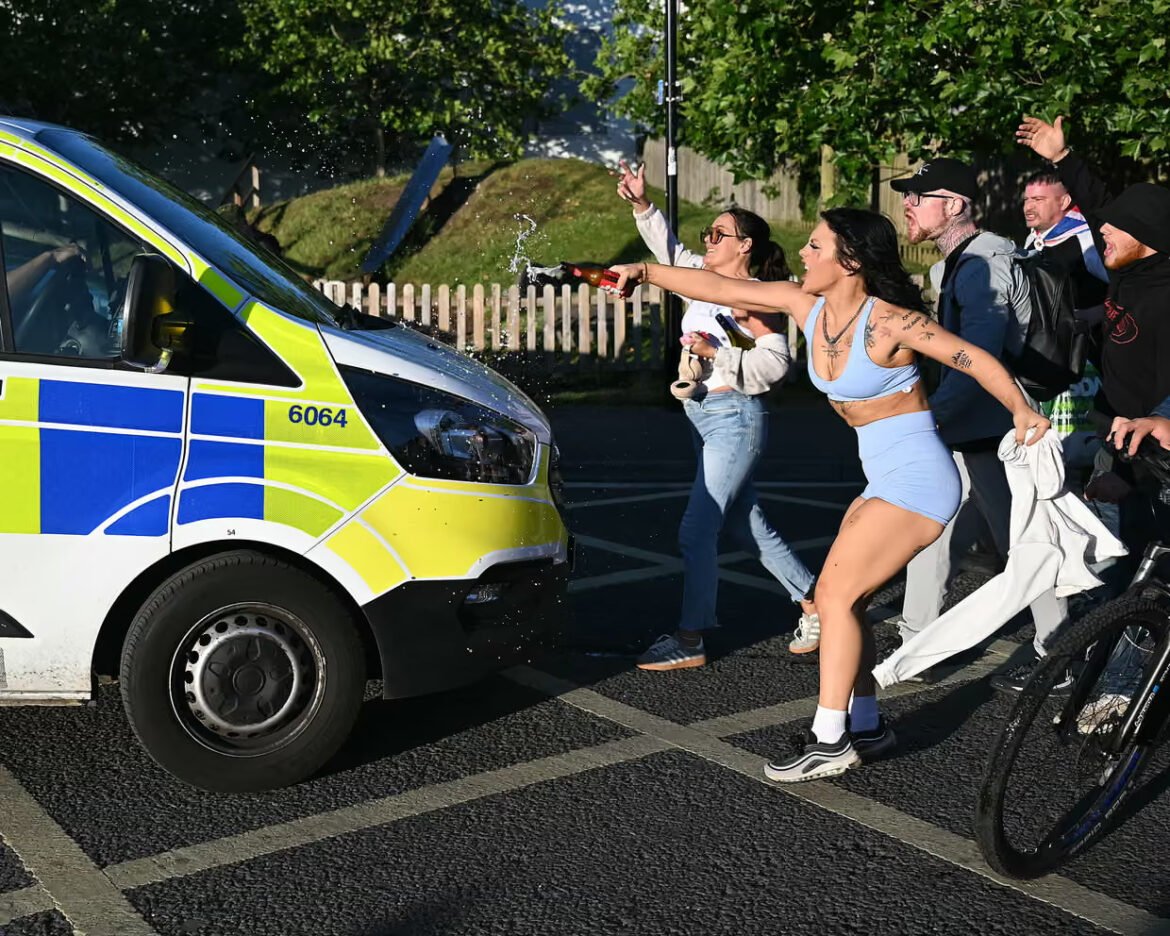 Group of people celebrate by spraying champagne toward a police van in a parking lot, with a woman in blue athletic wear leading the spray.
