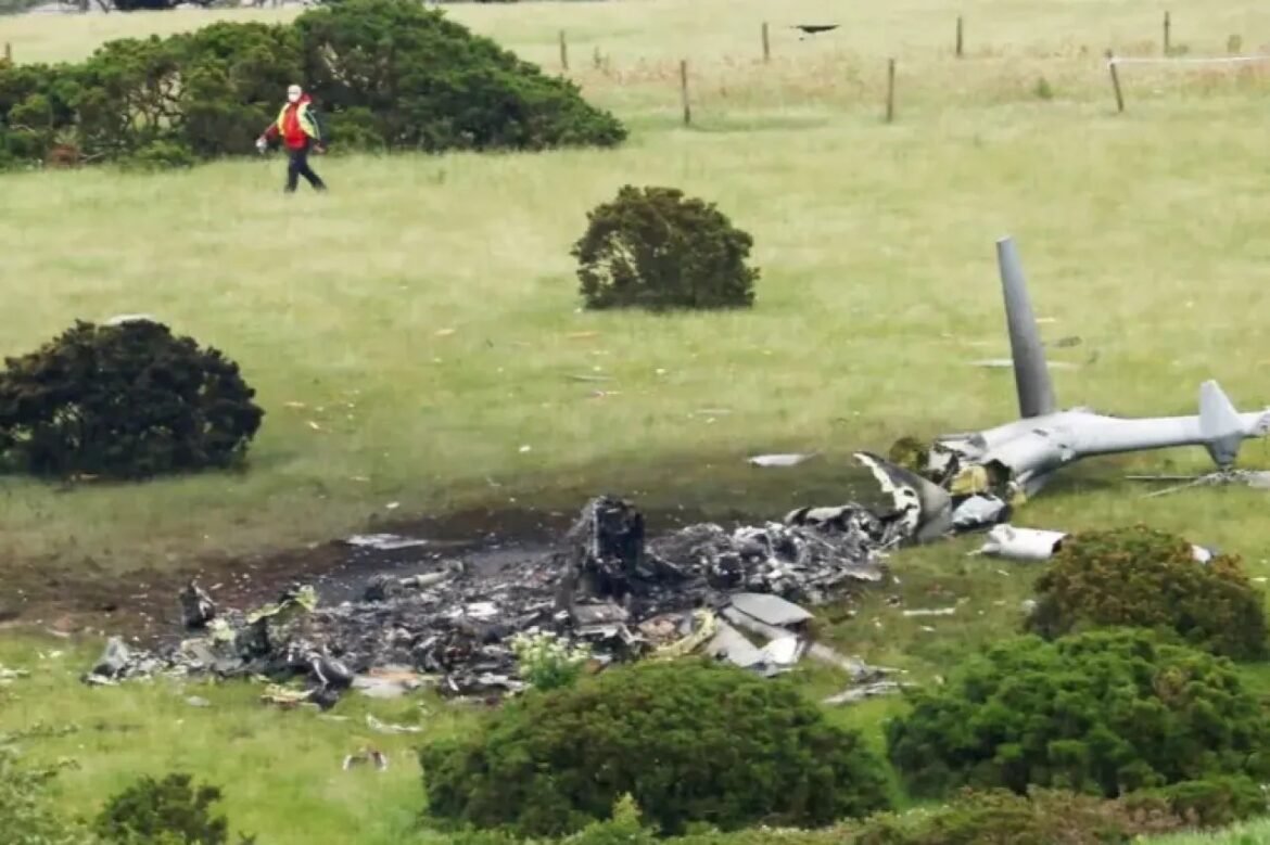 Wreckage of a small airplane in a grassy field with debris scattered; a person in a red jacket walks nearby as responders survey the site.