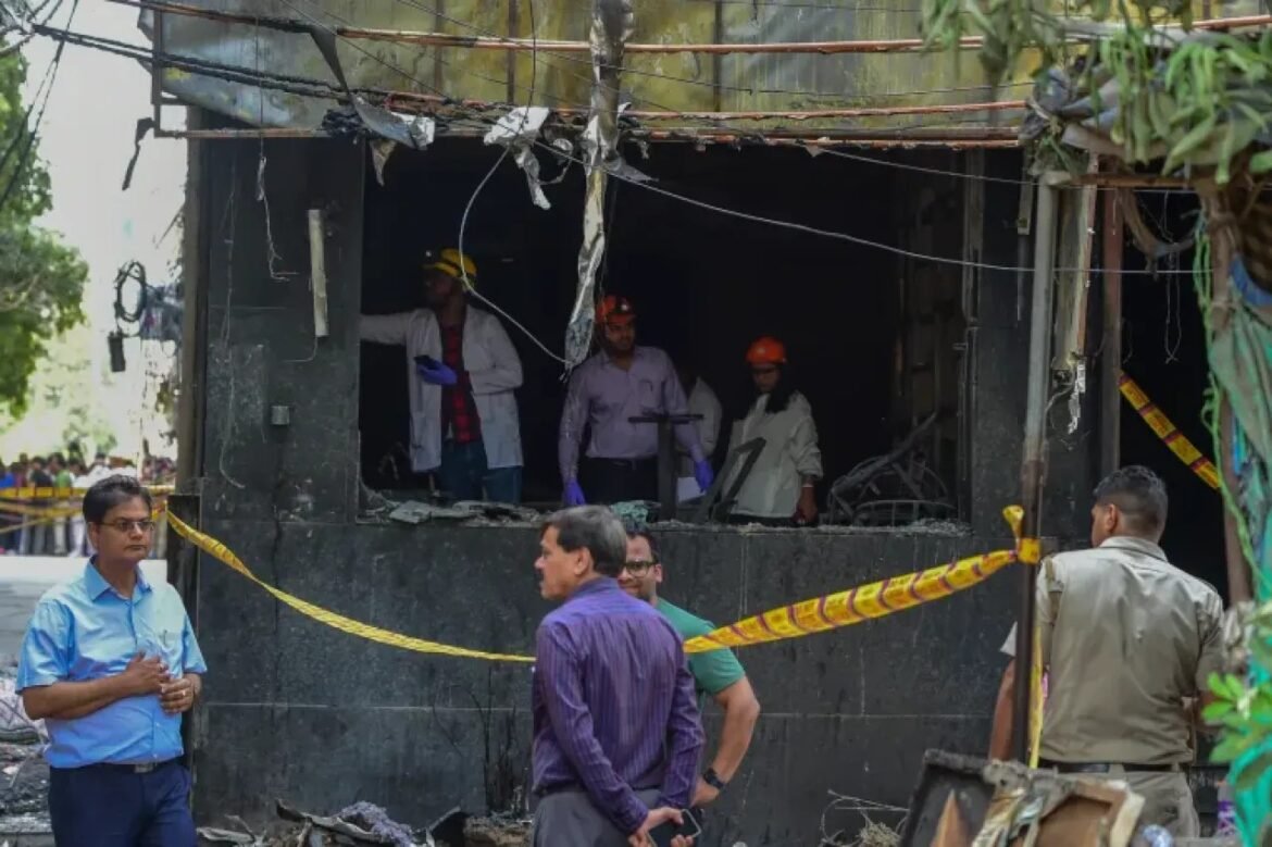 Group of men in helmets and safety gear inside a burned-out building, with yellow caution tape and onlookers outside the site.