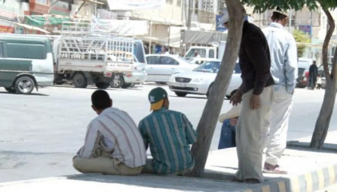 Two men sit on a curb by a busy street while a third stands nearby, near a tree and passing cars in the background.