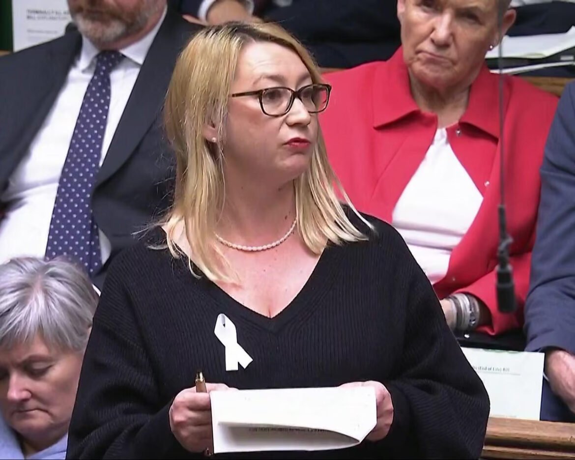 Woman with blonde hair and glasses speaks at a formal meeting, holding papers and a pen; wearing a white ribbon on a black sweater.