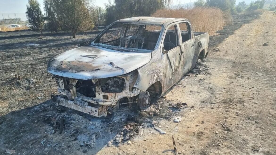 Charred, abandoned pickup truck with extensive fire damage sits in a dry, dirt field; front end gutted, wheels missing or damaged.
