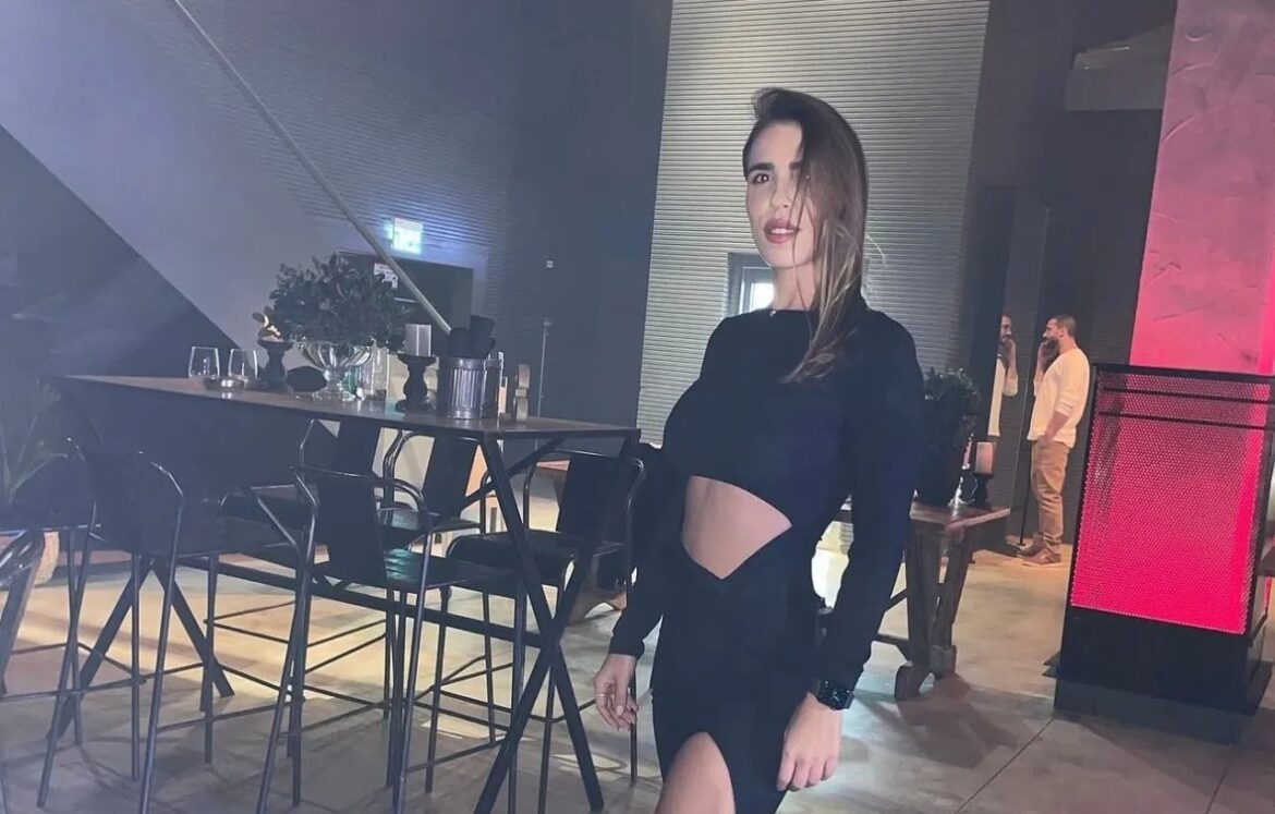 Woman in a black cut‑out dress standing beside a high table in a dimly lit event space, posing for the camera.
