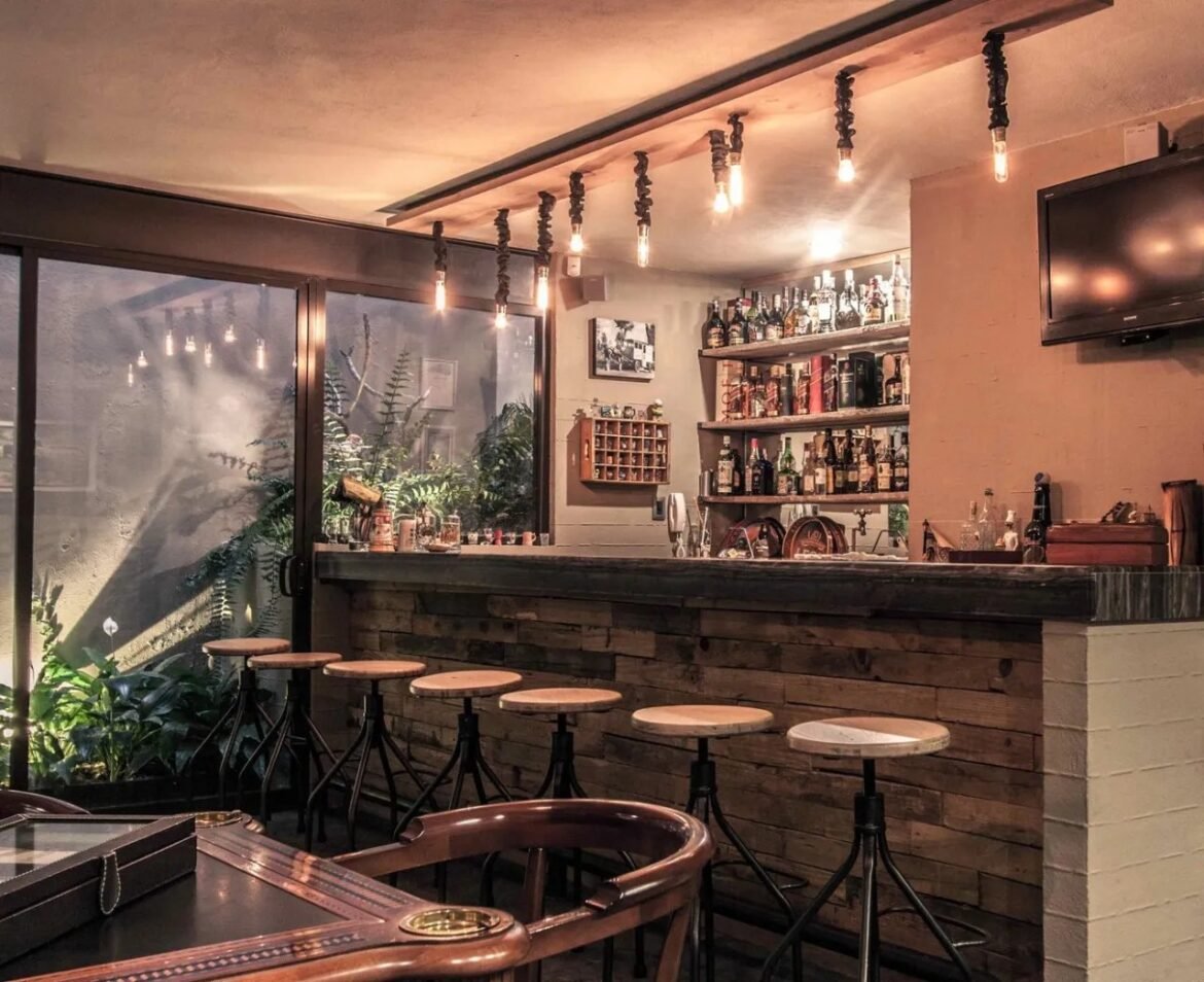 Cozy bar interior with a wooden counter, row of high stools, and bottles on wall shelves under warm hanging bulbs at dusk.