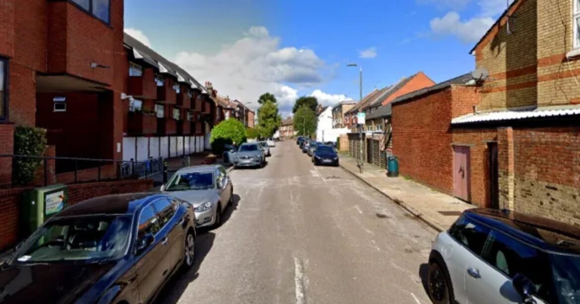 Residential street with cars parked along both sides, brick apartment blocks on the left and brick houses on the right, under a blue sky.