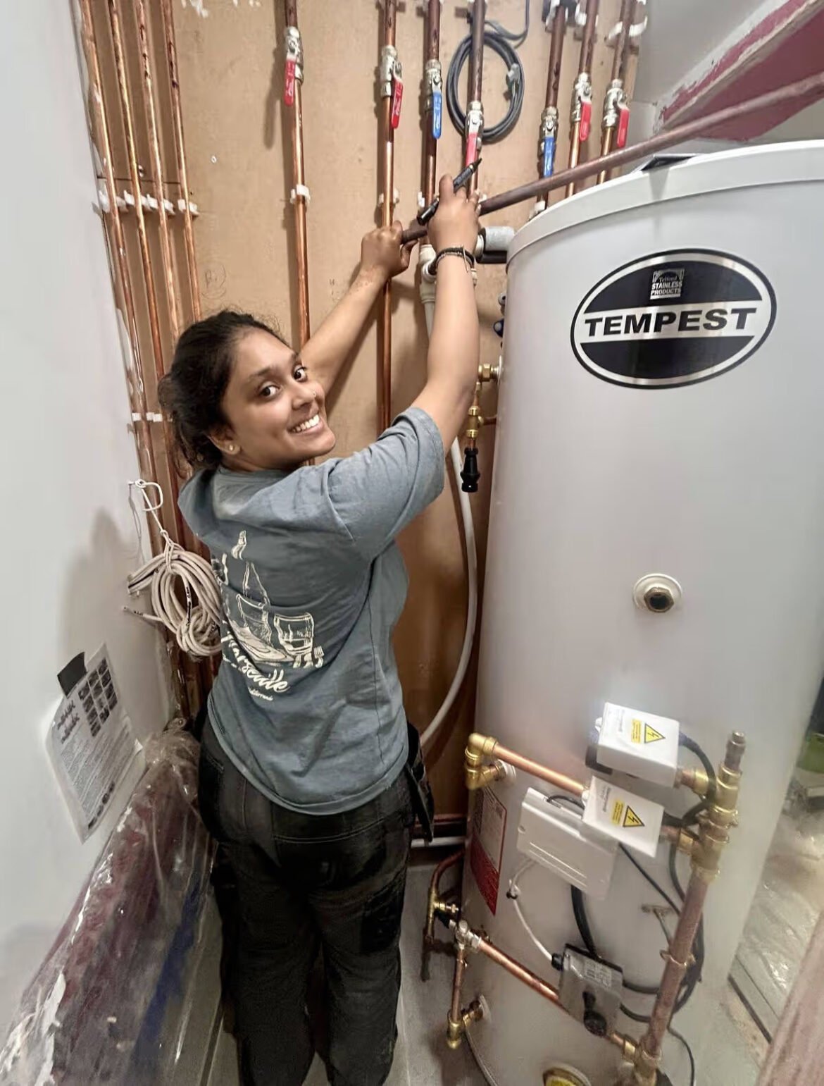Smiling woman repair technician tightening pipes beside a large white Tempest water heater in a utility room with copper plumbing.