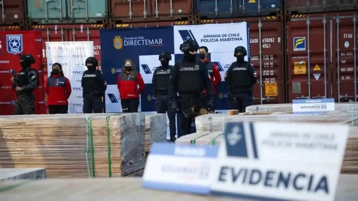 Group of armed officers in tactical gear standing behind pallets at a cargo dock, with an 'EVIDENCIA' sign in the foreground and shipping containers in the background.