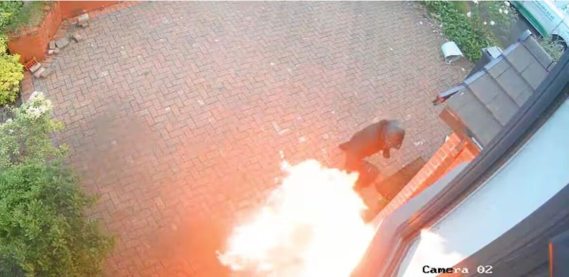Overhead security camera view of a bright explosion or large flame near a doorway on a brick patio, with a person standing to the right and greenery along the edge.