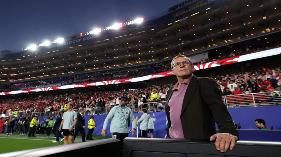Man in a purple shirt and dark blazer leaning on a railing at a crowded football stadium during a night match, foreground observer with stadium lights in the background.