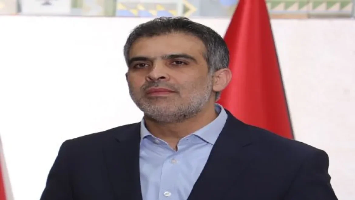 Portrait of a middle-aged man in a dark suit and light blue shirt, standing in front of a red flag.