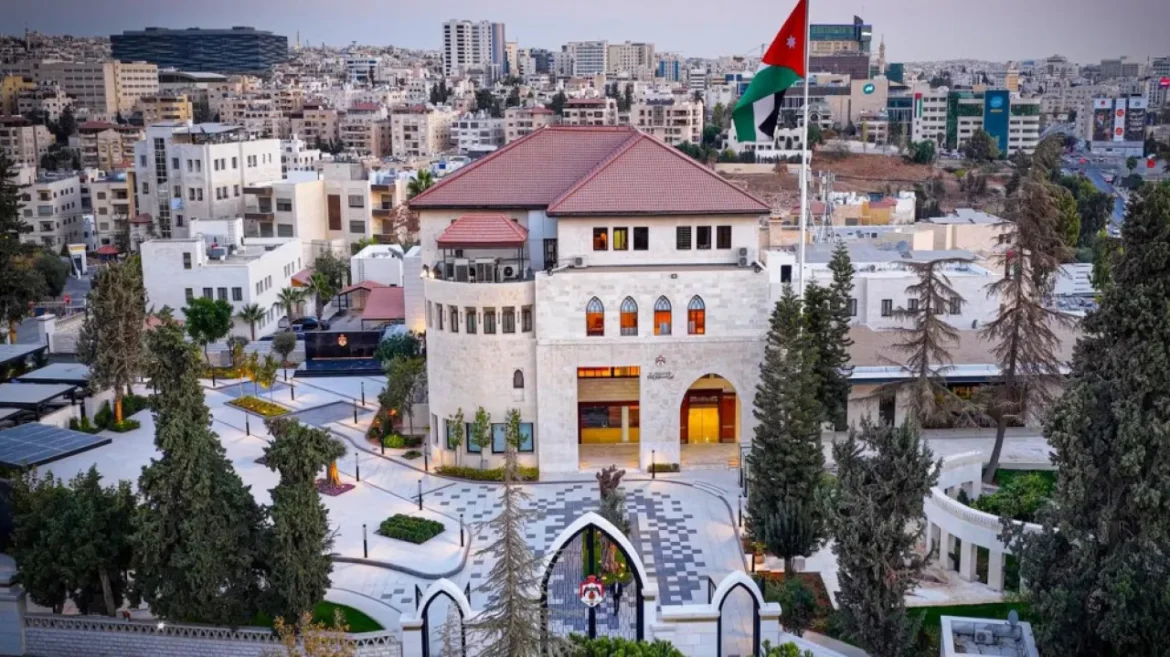 A white stone building with a red-tiled roof in a landscaped plaza, Jordan flag flying nearby, surrounded by trees and city streets.
