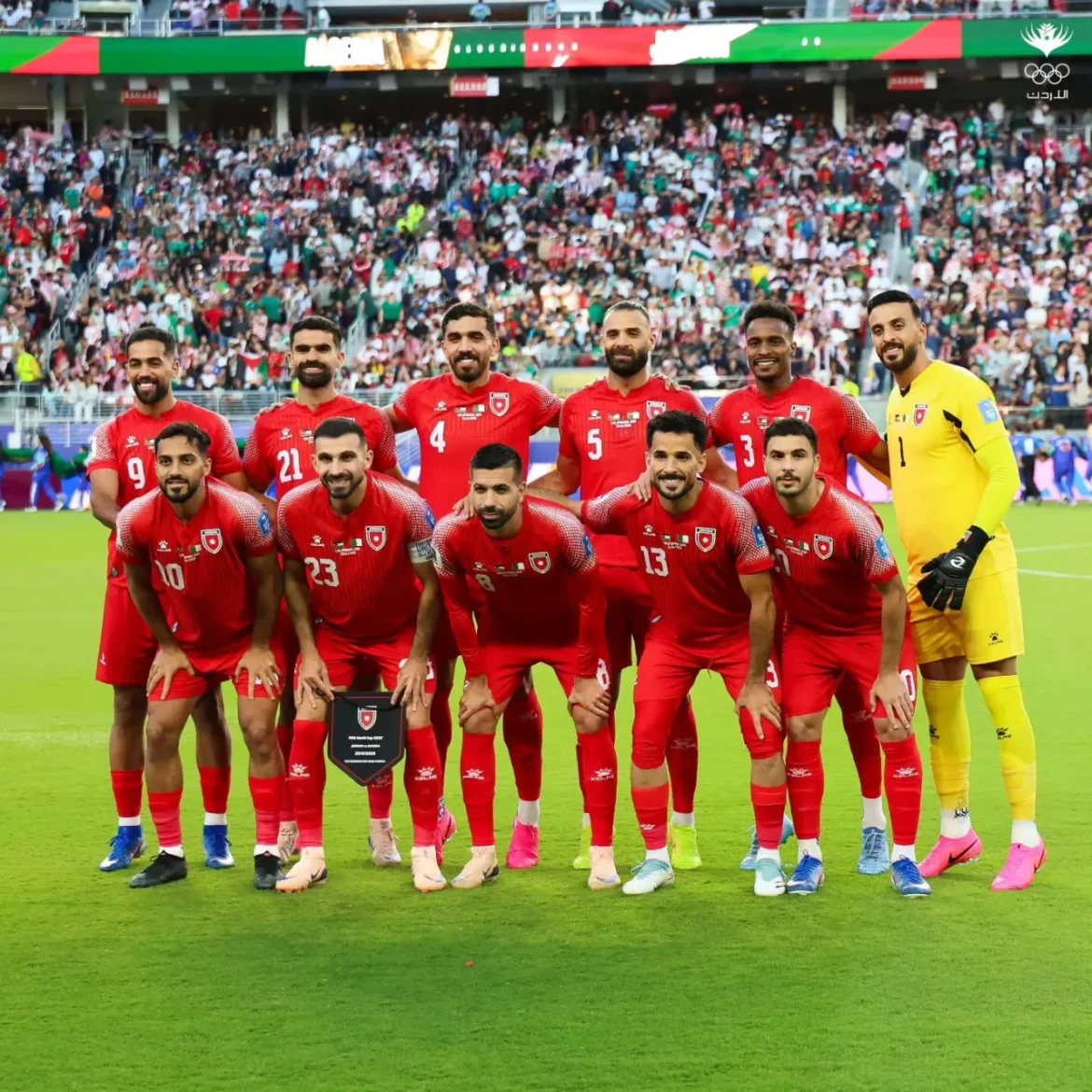 Twelve football players in red kits pose for a team photo on a grass pitch, with a yellow goalkeeper on the right, crowd-filled stands behind them and a trophy plaque visible on the ground.