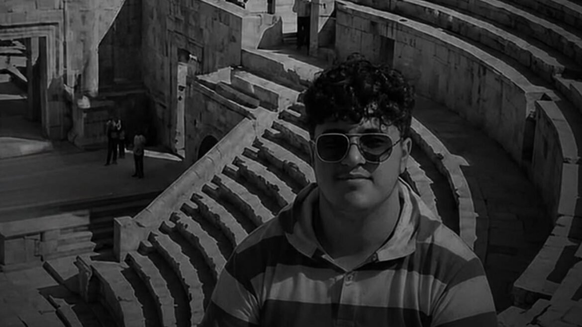 Man wearing sunglasses and a striped shirt stands in front of an ancient stone amphitheater backdrop.