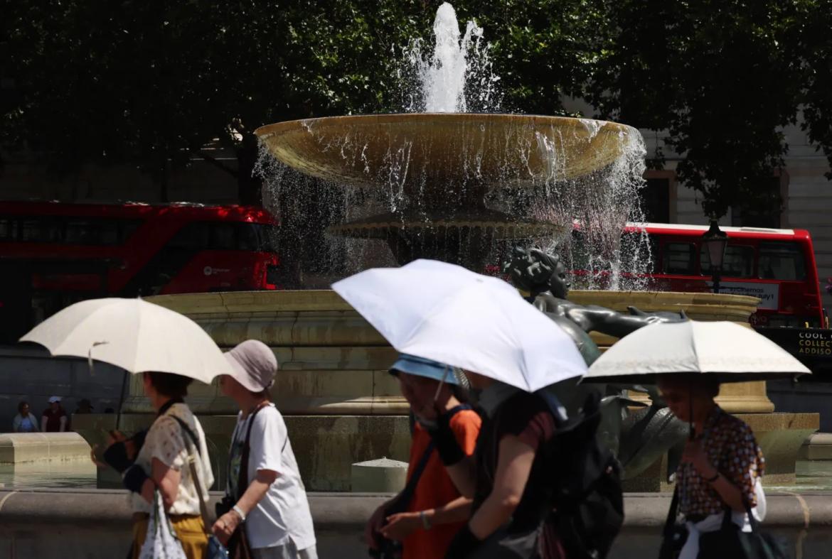 People holding umbrellas walk past a large stone fountain in a sunny city square, with red buses in the background.