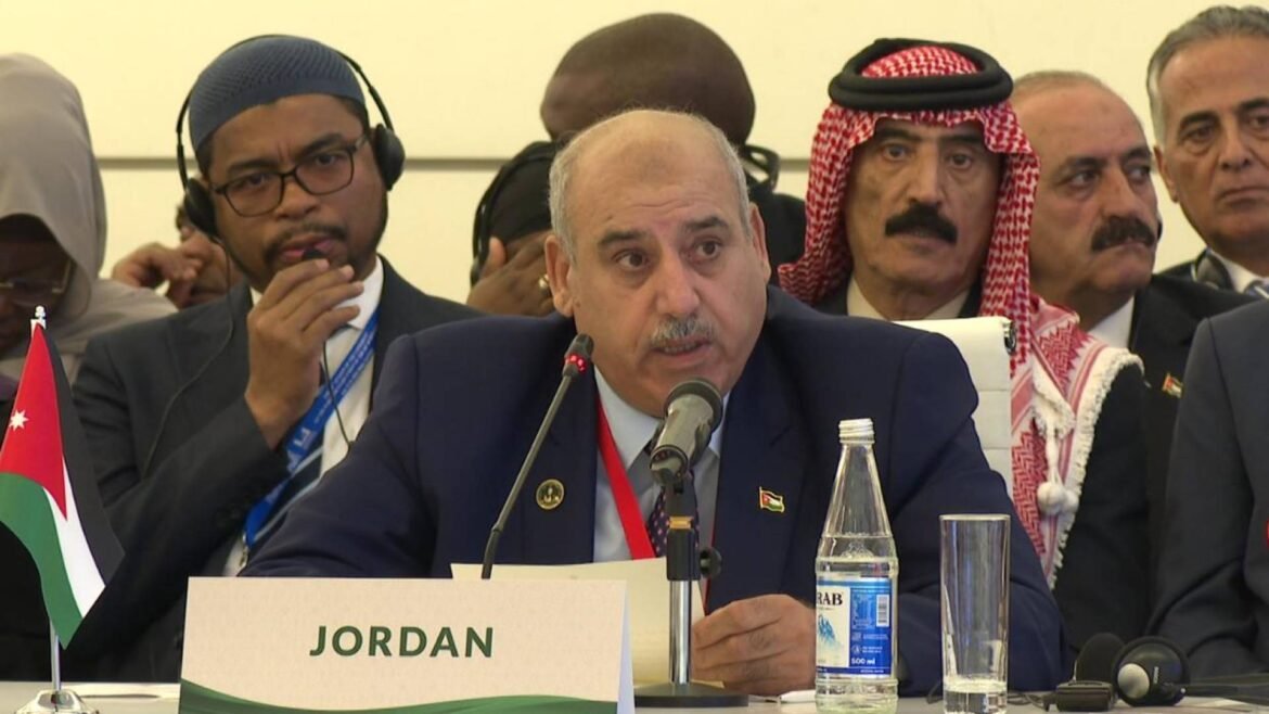 Jordanian official speaking at a conference, microphones in front, with a Jordan flag on the left and a water bottle nearby.