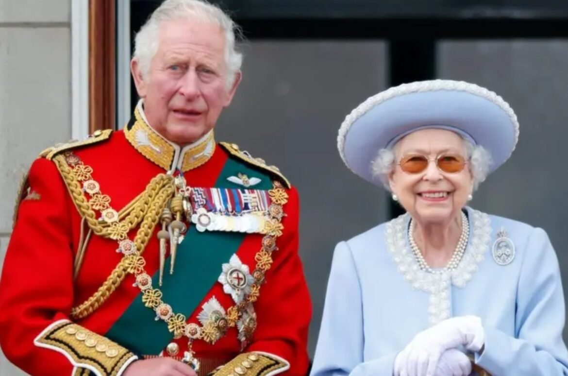 An elderly man in a red ceremonial military uniform with gold trim and many medals sits beside an elderly woman in a light blue outfit with a large white hat, smiling for the camera in a formal outdoor setting.
