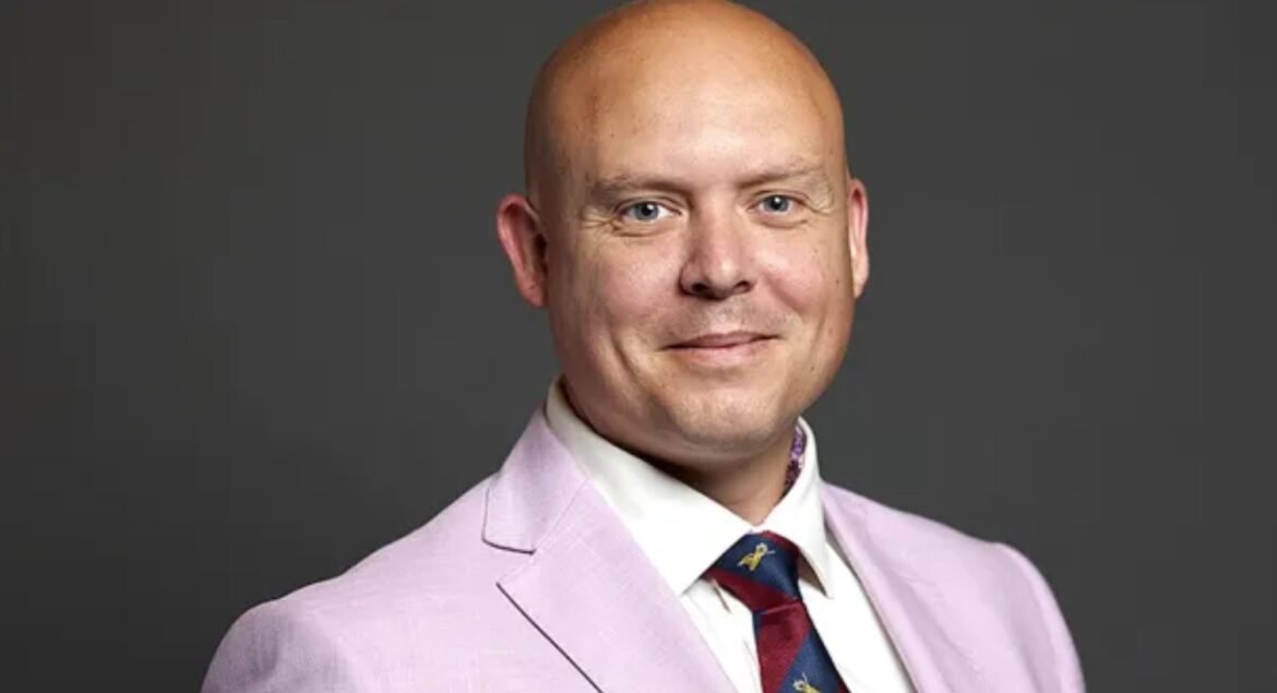 Portrait of a bald man in a light pink suit jacket and white shirt, smiling against a gray background.