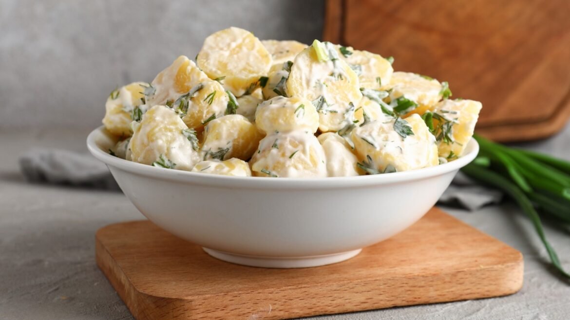 Creamy potato salad with dill and green onions in a white bowl on a wooden board