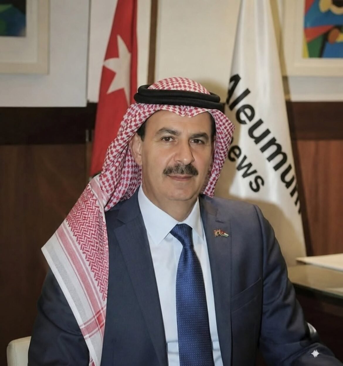 Man in a dark suit and blue tie wearing a red-and-white checkered keffiyeh, seated indoors with flags behind him.