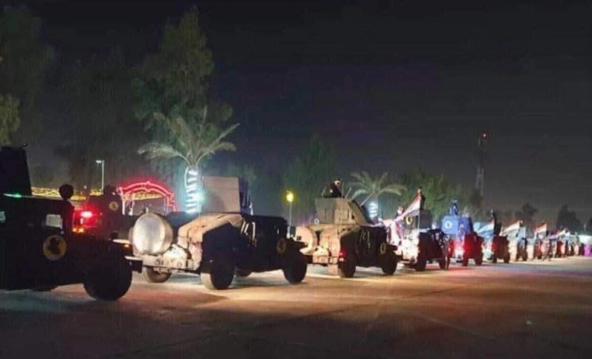 Line of military armored vehicles lined up on a dark street at night, with palm trees and decorative lights in the background.