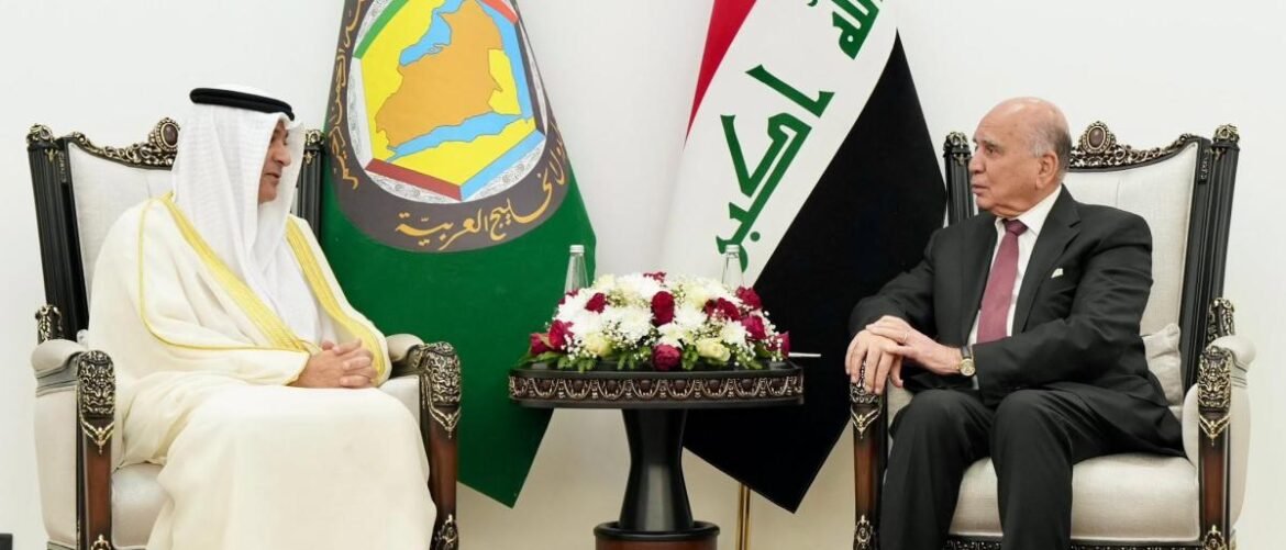 Two men in formal dress sit in ornate chairs across a small table with a flower centerpiece; UAE and Iraq flags in the background.