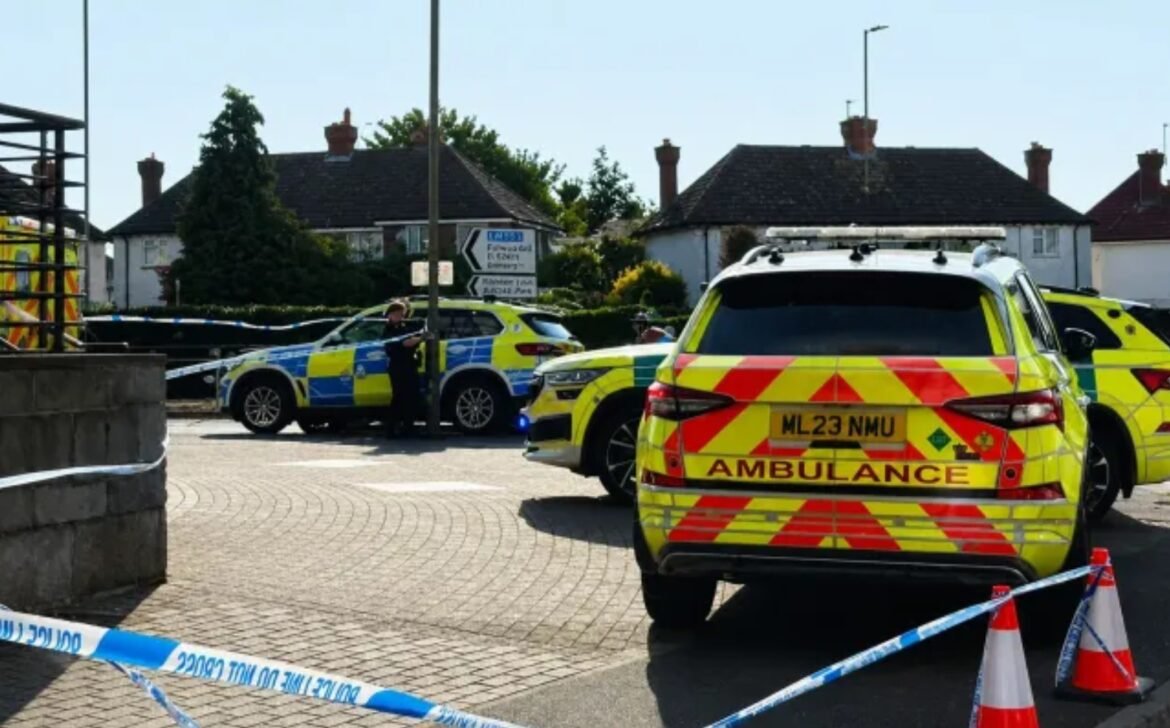 Emergency scene in a residential street with a yellow ambulance and police cars cordoned off by blue-and-white tape.