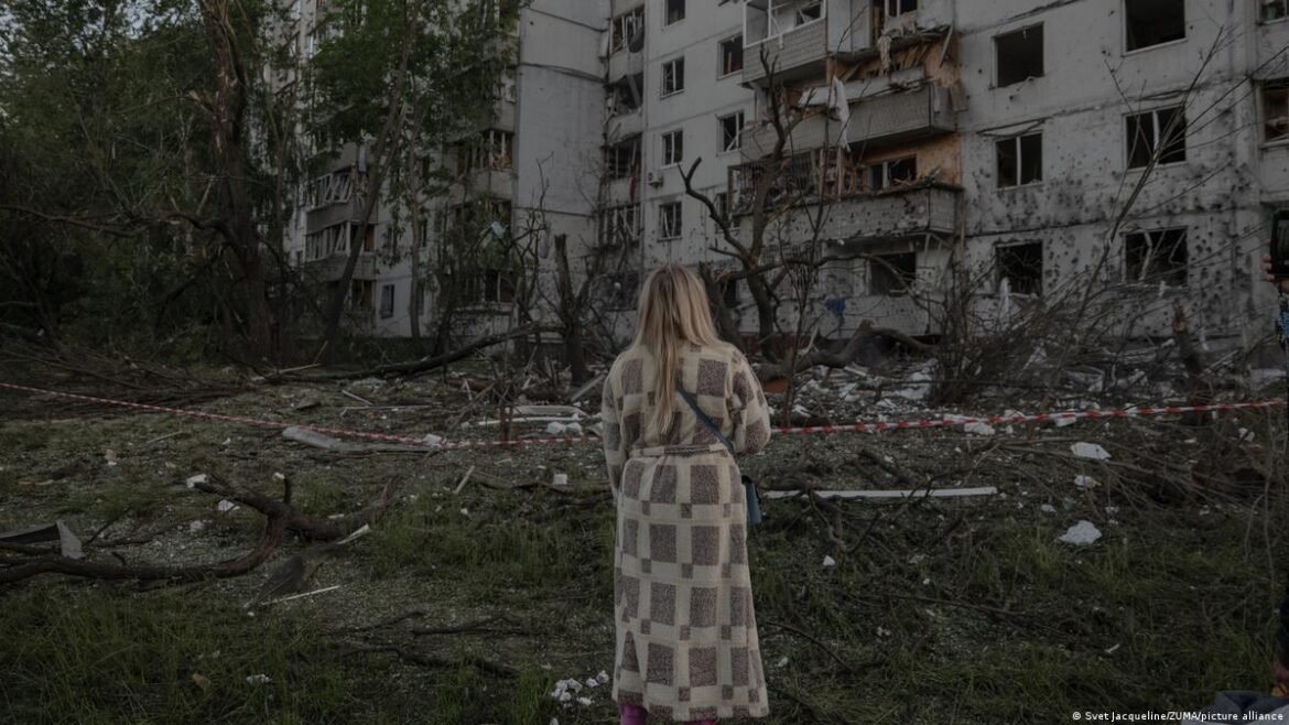 Woman in a checkered beige coat stands with her back to the camera, surveying a war-damaged apartment block and debris-scattered ground guarded by red tape.