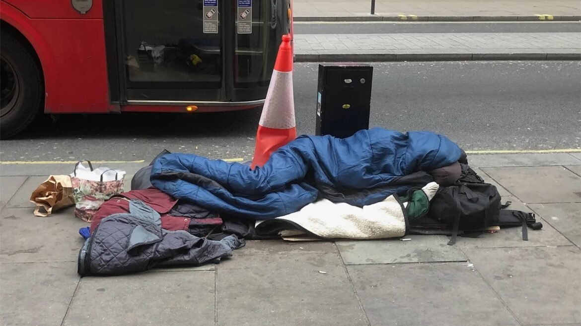 Person lying on the sidewalk under a pile of sleeping bags and coats, with bags nearby and a red bus, orange cone, and street in the background.