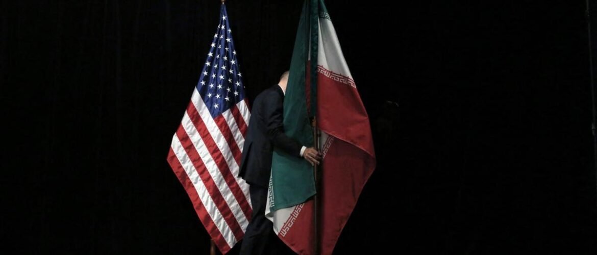Someone in a dark suit carries both the United States flag and the Iranian flag against a black background, suggesting a flag ceremony or formal event.
